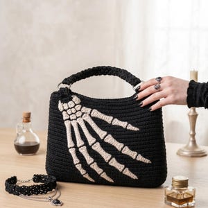 Peut inclure: Un sac à main en crochet noir orné d'un motif de main squelettique de couleur crème sur le devant. Le sac possède une anse tressée fixée par des anneaux en métal argenté. La texture est uniforme sur tout le corps en fil noir du sac.