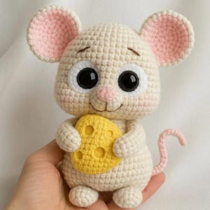 Puede incluir: Un juguete de ratón de ganchillo en crema, sosteniendo una cuña de queso amarillo. El ratón tiene grandes ojos negros, orejas rosas y una cola rosa. El juguete hecho a mano tiene un diseño caprichoso.