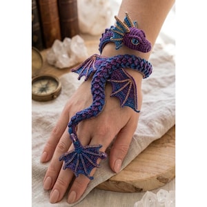 Op de afbeelding: Een gehaakte drakenarmband in paars en blauw die om de pols wikkelt en doorloopt naar een ring aan de vinger. De draak heeft gedetailleerde vleugels, een gestructureerd geschubd lichaam en een kop met blauwe ogen, gemaakt van paars en indigogaren.