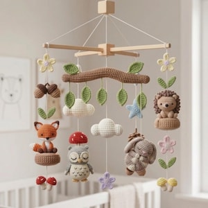 May include: A crochet baby mobile featuring a wooden frame with hanging yarn figures including a fox, owl, hedgehog, and rabbit. The mobile includes crocheted elements like green leaves, white clouds, red and white mushrooms, and pastel-colored flowers.