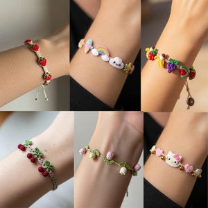 Pode incluir: Uma coleção de pulseiras de crochê feitas à mão. Cada pulseira apresenta designs exclusivos, incluindo morangos, um arco-íris, frutas, cerejas, flores e um design Hello Kitty. As pulseiras são de várias cores e são exibidas em um pulso.