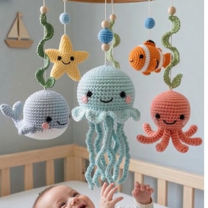May include: A handmade baby mobile featuring crocheted sea creatures: a whale, jellyfish, starfish, clownfish, and octopus. The mobile is suspended from a wooden ring with hanging strings. The sea creatures are in shades of blue, orange, yellow, and pink.