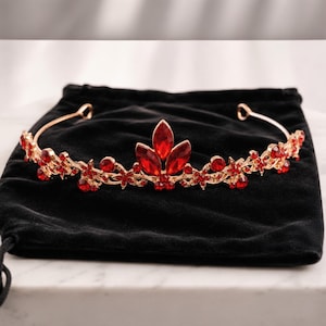 Op de afbeelding: Een goudkleurige tiara met rode edelstenen. De tiara heeft een centraal bloemmotief en kleinere bloemaccenten langs de band. Het rust op een zwarte fluwelen trekkoordtas, wat een opvallend contrast creëert.