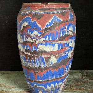 May include: A tall, decorative vase with a unique, layered design. The vase features a blend of red, blue, white, and black colors in a wavy pattern. The ceramic vase has a wide body and a slightly narrower opening at the top.