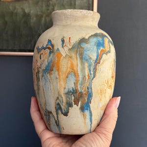 May include: A beige ceramic vase with a textured surface and a rounded body. The vase features a unique design with streaks of blue, orange, and brown paint. The vase is held in a hand.