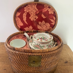 May include: A wicker basket with a lid, containing a small porcelain teapot and two cups. The basket is lined with red fabric with a floral pattern. The teapot and cups are decorated with floral designs in pink, green, and white.