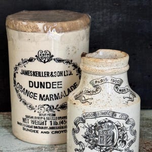 May include: A tall, cylindrical, off-white ceramic James Keiller & Son Ltd. Dundee Orange Marmalade jar with a paper-covered lid, next to a smaller, speckled off-white ceramic Grey-Poupon Dijon mustard jar featuring black printed text and graphics.