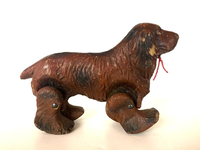 May include: A brown wooden toy dog with a red string attached to its neck. The dog has four wheels and is a vintage toy.