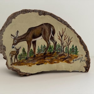 May include: Hand-painted artwork on a piece of wood bark. The image depicts a deer and a fawn in a forest setting, with trees and foliage. The artwork is signed "Sonny" with the year "06".