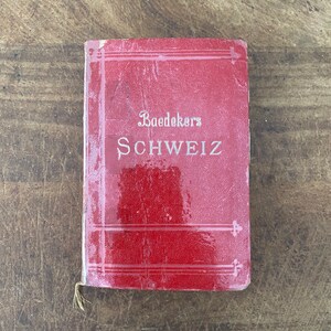 May include: A vintage red book titled "Baedekers SCHWEIZ" with gold lettering. The book has a textured cover and a small, frayed tassel. The book is lying on a wooden surface.