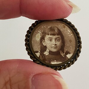 May include: A small, round, brass locket with a braided rope design around the edge. Inside the locket is a black and white photograph of a young girl with a serious expression.
