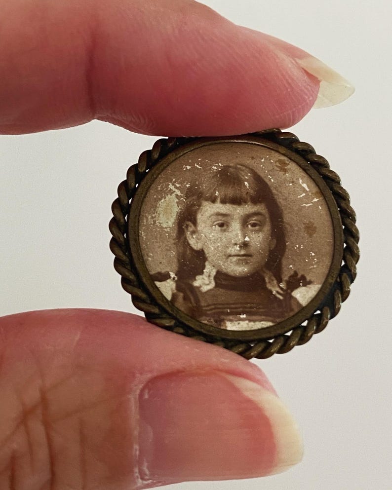 May include: A small, round, brass locket with a braided rope design around the edge. Inside the locket is a black and white photograph of a young girl with a serious expression.