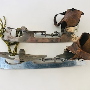 May include: A pair of vintage ice skate blades with leather straps. The blades are silver and have a worn look. The straps are brown leather and have a buckle closure.