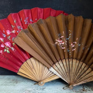 May include: Two antique hand fans. One is red with floral designs in pink, white, and blue. The other is brown with similar floral accents. Both fans have wooden handles and are partially open, showcasing their pleated design.
