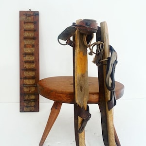 May include: A pair of antique wooden ice skates with leather straps and buckles. The skates are standing on a wooden stool with three legs. A wooden sign with handwritten text is in the background.