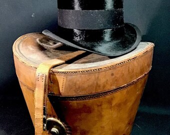 May include: A brown leather hat box with a strap and buckle. A black top hat sits on top of the box.