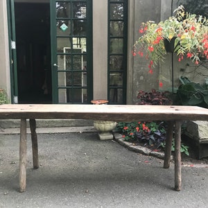 May include: A rustic wooden table with a long, rectangular top and four sturdy legs. The table is made of rough-hewn wood and has a natural, weathered finish. It is sitting on a paved patio in front of a building with green doors.