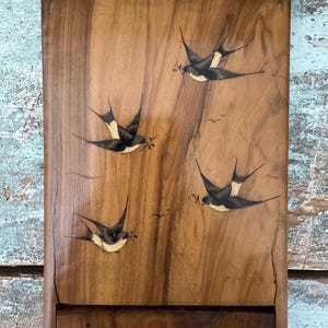 May include: Wooden box with a hinged lid, featuring a detailed illustration of swallows in flight. The swallows are depicted in black and white, carrying twigs, set against a warm, wood-grain background. The box has a rectangular shape.