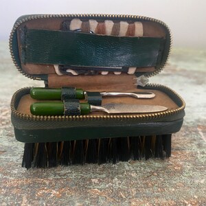 May include: A green leather travel case with a zipper closure. The case contains a small brush and two tools with green handles. The case is open and the tools are visible.