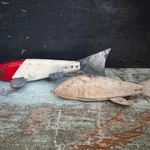 May include: Two wooden fish sculptures. One is painted red and white with a metal tail and fins. The other is a natural, weathered wood color. Both are on a distressed, painted surface.