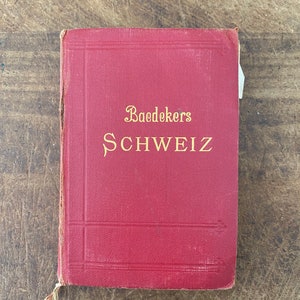 May include: A vintage red book titled "Baedekers SCHWEIZ" in gold lettering. The book has a textured cover and shows signs of wear, suggesting it is an antique travel guide. The book is resting on a wooden surface.