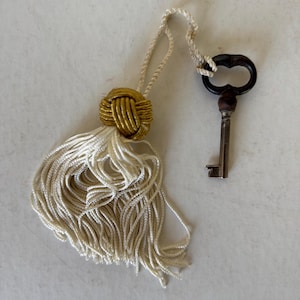 May include: A decorative tassel with a gold-colored knot and cream-colored fringe, next to a vintage key with a dark brown bow-shaped handle. The key is made of silver-colored metal. The tassel and key are on a white background.