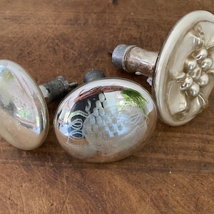 May include: Three antique glass door knobs with silver finish. Two knobs have a round shape with a raised design, and one knob has a flat, circular shape with a floral design.