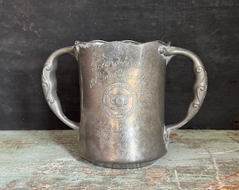 May include: Antique silver-colored metal tankard with two ornate handles. The tankard has a scalloped rim and engraved text and a circular emblem on the front. The text reads "Tennis Prize 120 Yards".