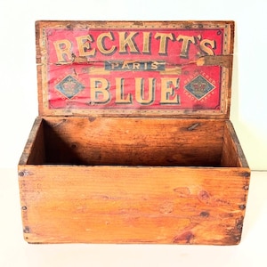 May include: An open, antique wooden box with a red and gold label on the lid. The label reads "RECKITT'S PARIS BLUE" in gold lettering. The box is made of light brown wood and shows signs of age.