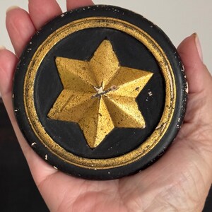 May include: A circular decorative object featuring a raised, six-pointed gold star centered on a matte black background. The outer edge of the circle is finished with a matching gold border, and the black surface shows signs of distressed wear.