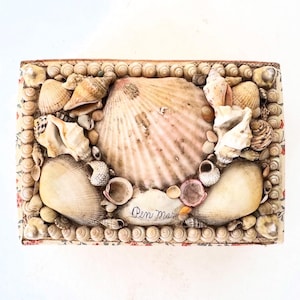 May include: A small, rectangular box covered in a floral fabric with a border of small seashells. The box is filled with a variety of seashells, including a large scallop shell in the center. The text "Oen Mar" is written on the box.