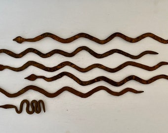 May include: A set of seven rusty metal snake sculptures, arranged in a wavy pattern on a white background. The snakes are all the same size and shape, and they are all facing the same direction. The smallest snake is curled up at the bottom of the arrangement.