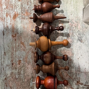 May include: A selection of wooden finials in diverse shapes and shades of brown, arranged vertically. The finials have screw threads for attachment and are displayed against a weathered, distressed wooden background.
