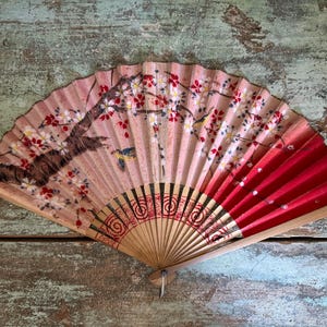 May include: Hand-painted folding fan with a floral design. The fan features a light pink background with a branch of a tree with red and white flowers. The right side of the fan is red. The fan is made of wood and paper.