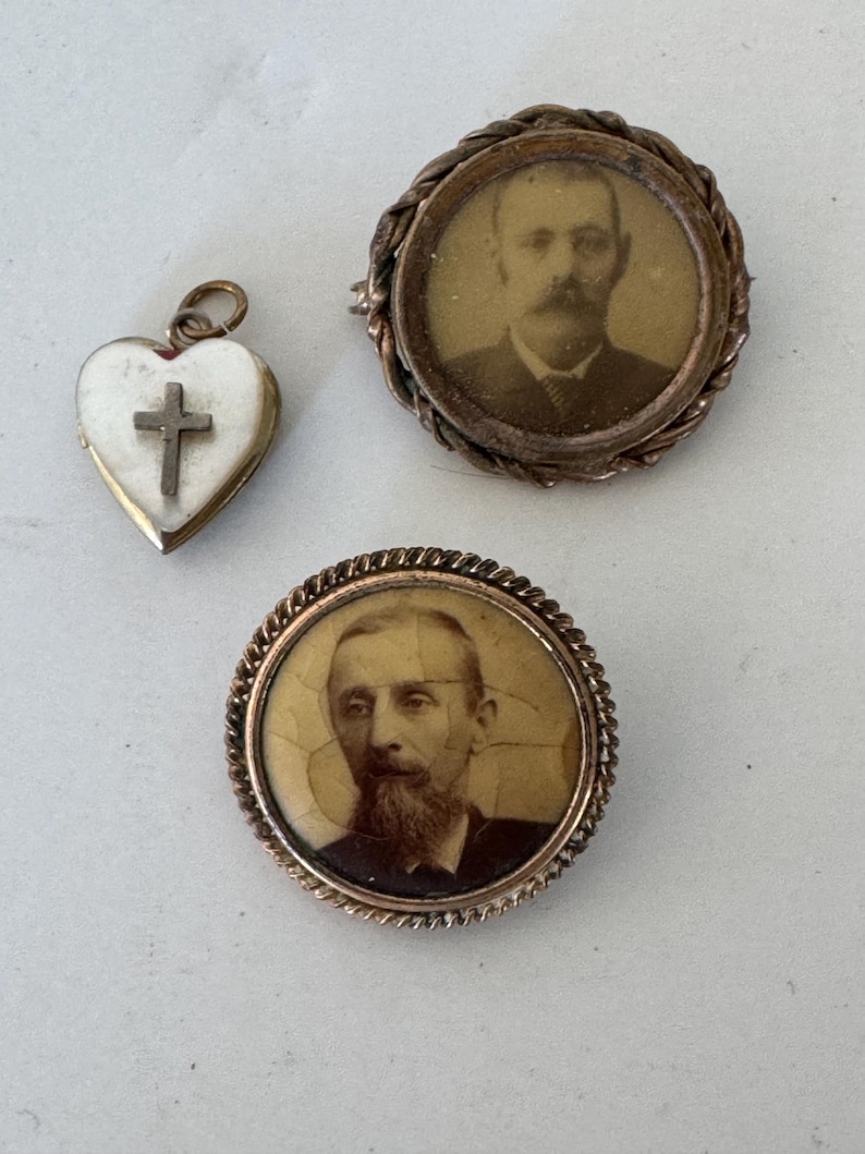 May include: Three antique gold-toned brooches. Two of the brooches feature a portrait of a man in a round frame. The third brooch is a heart-shaped locket with a white cross.