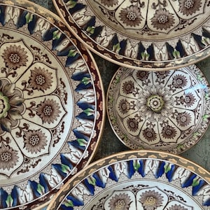 May include: Overhead shot of a collection of antique ceramic plates. The plates feature a floral design in brown, blue, and green on a cream background. The plates vary in size and have a decorative border.