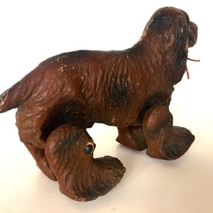 May include: A brown wooden toy dog with a red string attached to its mouth. The dog has four legs and is standing on a white surface.