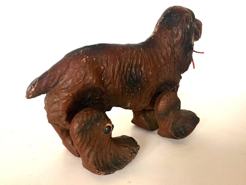 May include: A brown wooden toy dog with a red string attached to its mouth. The dog has four legs and is standing on a white surface.