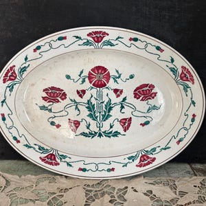 May include: Oval serving platter with a white background and a floral design. The design features red poppies and green leaves, with a border of smaller red flowers and green vines. The platter is likely made of ceramic or porcelain.