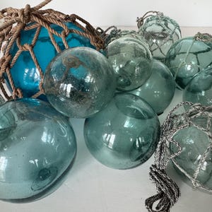 May include: A group of blue and green glass fishing floats, some with a net covering, are arranged on a white surface.