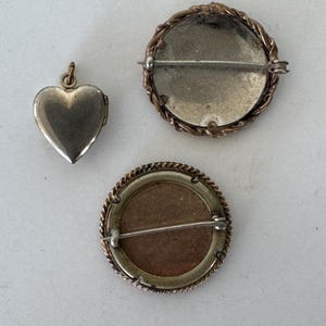 May include: Three vintage brooches, one heart-shaped locket and two round brooches with a braided metal design around the edges. The brooches are silver-toned with a gold-toned metal backing.