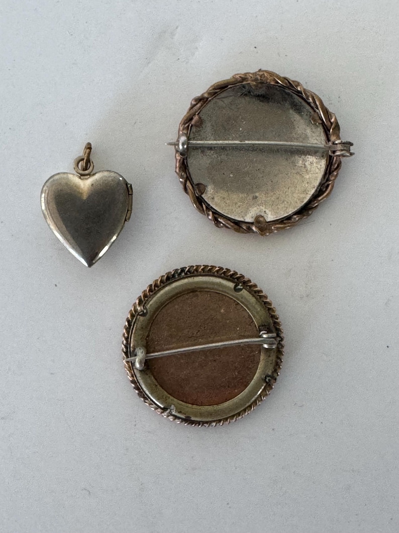 May include: Three vintage brooches, one heart-shaped locket and two round brooches with a braided metal design around the edges. The brooches are silver-toned with a gold-toned metal backing.