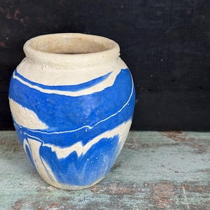 May include: A small, handmade ceramic vase with a rounded body and a wide opening. The vase features a striking design of blue and white swirls, creating a marbled effect. The exterior has a textured, matte finish, and the vase sits on a weathered wooden surface.