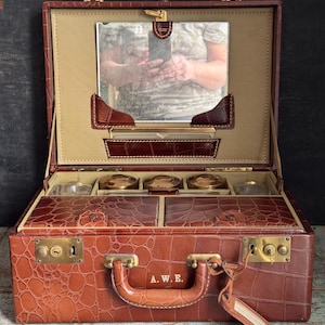 May include: Open brown leather travel case with a crocodile pattern. The case has a handle, brass hardware, and a mirror inside the lid. The interior is tan with brown leather accents. The initials "A.W.E." are on the handle.