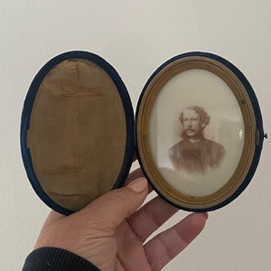May include: An antique oval portrait in a velvet-lined case. The portrait is sepia-toned, featuring a man with a mustache and curly hair. The case is open, revealing the portrait and the blue velvet lining.