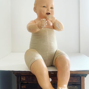 May include: A vintage porcelain baby doll with a painted face and open mouth, wearing a beige cloth diaper. The doll is sitting on a wooden surface with drawers.