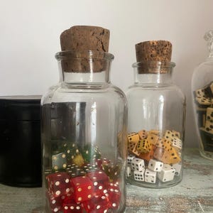 May include: Two glass jars with cork lids filled with colorful dice. The jars are on a wooden surface.
