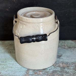 May include: A cylindrical off-white stoneware crock featuring a matching lid and a wire bail handle with a black wooden grip. The container has a smooth, glazed finish and a sturdy base, designed for storage with a simple, functional aesthetic.
