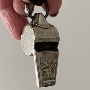 May include: A silver-colored whistle with a loop for attaching to a chain. The text "THE ACME THUNDERER MADE IN ENGLAND" is visible on the front. The whistle is held by a person.