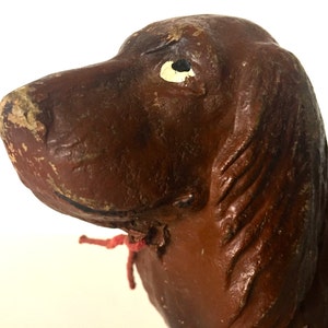 May include: Close-up of a brown ceramic dog figurine with a white eye. The dog has a red ribbon tied around its neck.